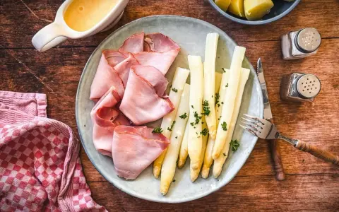 Spargel mit Schinken, garniert mit Petersilie, auf einem Teller mit Sauce Hollandaise und Kartoffeln daneben.