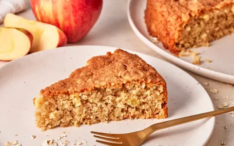 Ein saftiges Stück Apfel Haferflocken Kuchen liegt auf einem Teller, daneben ein ganzer Apfel und Haferflocken.