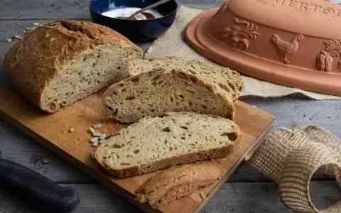 Frisch gebackenes Dinkelbrot mit Körnern, aufgeschnitten auf einem Holzbrett. Daneben ein Römertopf, eine Schüssel mit Joghurt und ein Messer.