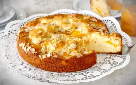 Ein goldbrauner Landfrauen Aprikosenkuchen, bestreut mit Mandelblättchen, wird auf einer zarten Tortenplatte serviert. Ein Stück wird gerade herausgehoben.