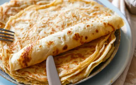 Ein Stapel goldbrauner Pfannkuchen, einer davon aufgerollt, mit Messer und Gabel. Perfekt für ein leckeres Pfannkuchen Rezept!