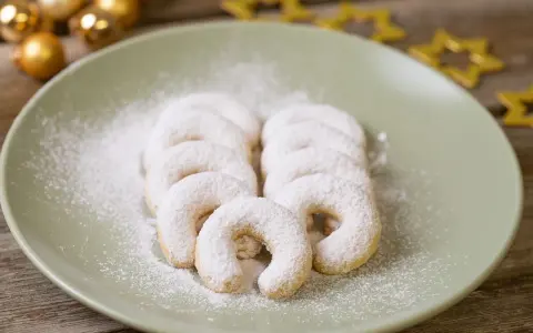 Vanillekipferl, bestäubt mit Puderzucker, liegen auf einem Teller. Goldene Kugeln und Sterne im Hintergrund deuten auf Weihnachten hin.