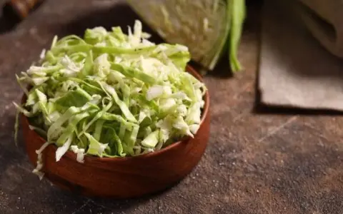 Fein geschnittener Spitzkohl in einer Holzschale, bereit, in der Pfanne gebraten zu werden. Ein halber Spitzkohl liegt im Hintergrund.