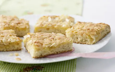 Saftiger mandelkuchen vom blech, goldbraun gebacken und mit knusprigen Mandelblättchen bestreut, liegt auf einer weißen Platte.