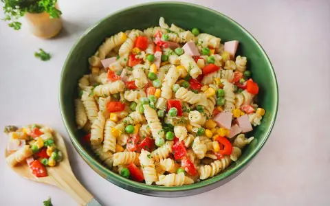 Ein bunter Nudelsalat mit Fleischwurst, Erbsen, Mais und Paprika in einer grünen Schüssel.