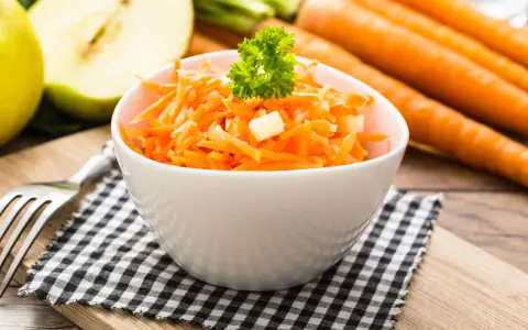 Frischer Karotten-Apfel-Salat in einer weißen Schüssel, garniert mit Petersilie. Im Hintergrund liegen ganze Karotten und ein halber Apfel.