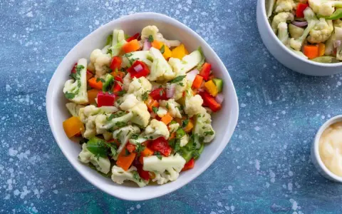 Frischer blumenkohl salat mit Paprika, Karotten und Kräutern, serviert in einer weißen Schüssel. Daneben eine kleine Schale mit Dip.