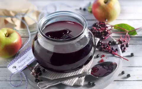 Ein Glas mit dunklem Holunder Apfel Gelee, daneben Äpfel und Holunderbeeren. Ein Löffel mit Gelee liegt bereit.