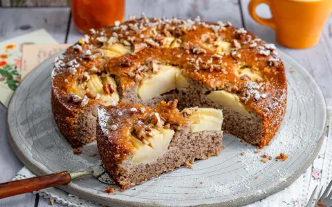 Ein saftiger Apfel-Birnen-Kuchen, bestreut mit Nüssen und Puderzucker, wird auf einem Teller serviert. Ein Stück ist bereits herausgeschnitten.