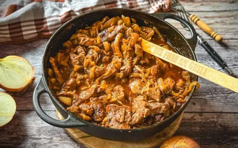 Herzhaftes Zwiebelfleisch-Rezept in Gusseisenpfanne, mit Zwiebeln und Fleischstücken in einer würzigen Sauce. Ein Holzlöffel mit der Aufschrift "Die Frau am Grill" liegt darin.