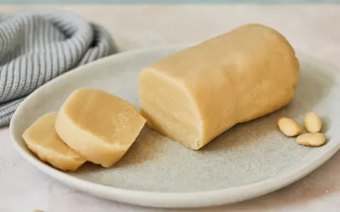 Frisch zubereitetes Marzipan zum **Marzipan selbst machen**. Ein Block und zwei Scheiben liegen auf einem Teller, daneben Mandeln.