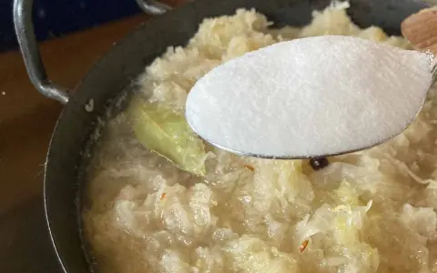 Zucker wird zu köchelndem Sauerkraut nach Omas Rezept gegeben. Ein Lorbeerblatt schwimmt im Topf.