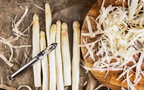 Frischer weißer Spargel wird geschält, die Spargelschalen liegen daneben. Ein Sparschäler liegt auf den Spargelstangen.