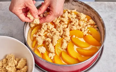Hände verteilen Streusel auf einem Pfirsichkuchen in einer Springform.