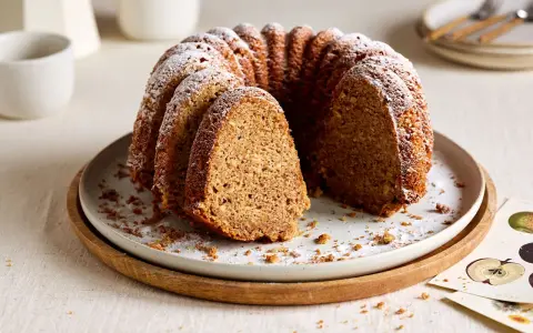 Ein saftiger Kuchen mit Dinkelmehl, bestäubt mit Puderzucker, liegt auf einem Teller. Ein Stück ist herausgeschnitten.