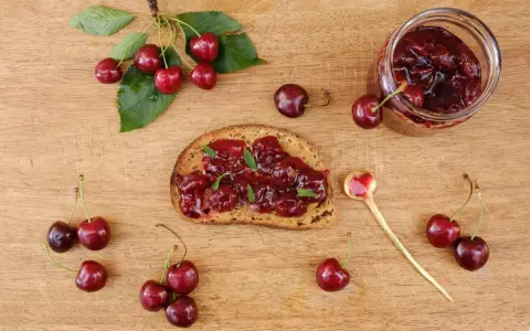 Frisches Brot mit Kirschmarmelade, garniert mit Minze. Ein Glas Kirschmarmelade und frische Kirschen daneben. Perfekt für ein Kirschmarmelade Rezept.