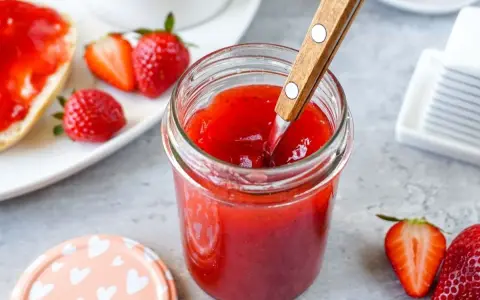 Ein Glas mit leuchtend roter Erdbeermarmelade, die mit einem Holzlöffel umgerührt wird. Daneben frische Erdbeeren und ein Stück Brot mit Marmelade – ein Genuss mit Pfiff!