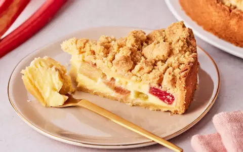 Ein Stück von Omas Rhabarberkuchen mit Pudding und Streuseln auf einem Teller, daneben ein goldener Löffel und rote Rhabarberstangen.