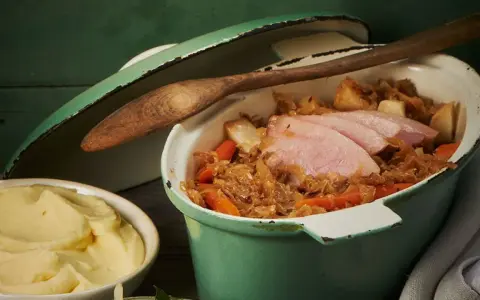 Herzhafter Szegediner Gulasch mit zarten Fleischscheiben, Sauerkraut und Karotten in einem grünen Topf, daneben eine Schüssel cremiges Kartoffelpüree.