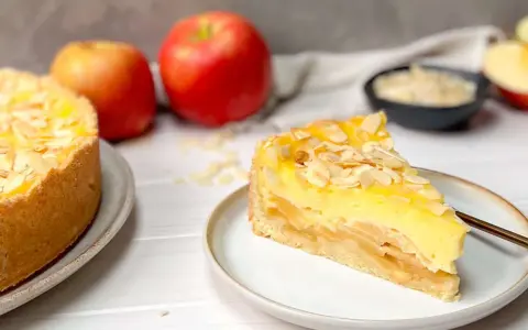 Ein saftiger Apfelkuchen mit Schmandguss, bestreut mit Mandelblättchen. Daneben liegen frische Äpfel.