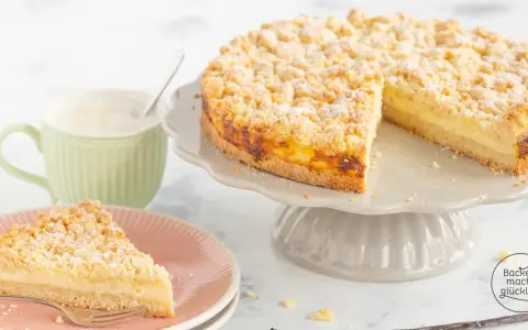 Ein saftiger Streuselkuchen mit Puddingfüllung, bestäubt mit Puderzucker. Daneben eine Tasse Cappuccino.