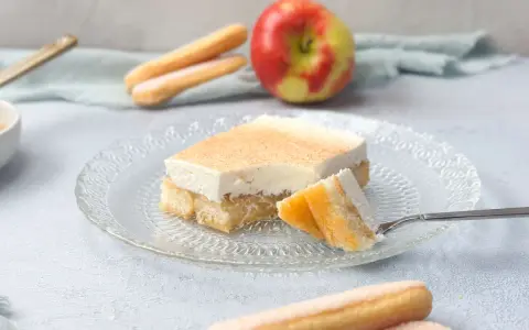 Ein Stück Apfel-Schmand-Kuchen aus der Springform, mit Löffelbiskuits und einem Apfel im Hintergrund.