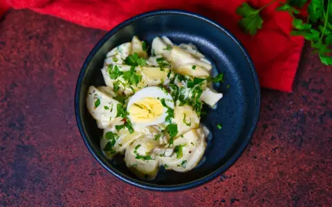 Ein cremiger Kartoffelsalat mit Mayo, garniert mit Petersilie und einem halben Ei, serviert in einer dunklen Schüssel.