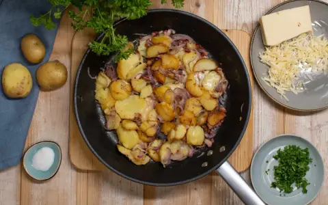 Bratkartoffeln mit Zwiebeln und Käse in der Pfanne, dazu frische Petersilie und Salz.