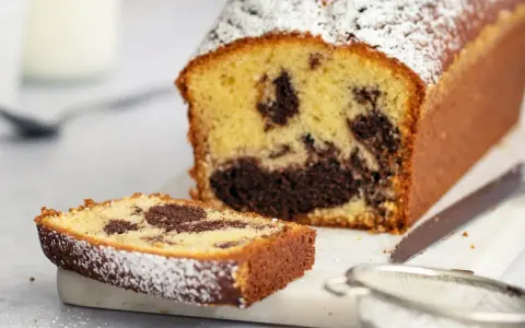 Ein saftiger marmorkuchen mit Puderzucker bestäubt, daneben ein Stück Kuchen und ein Sieb. Perfekt mit einem Klecks Sahne.