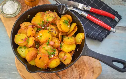 Bratkartoffeln mit Zwiebeln und Speck in einer Gusseisenpfanne, garniert mit Schnittlauch. Dazu grobes Salz und ein Messer-Gabel-Set.