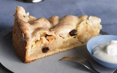 Ein Stück gedeckter Apfelkuchen mit Rosinen und Zuckerguss, serviert mit einem Klecks Sahne.