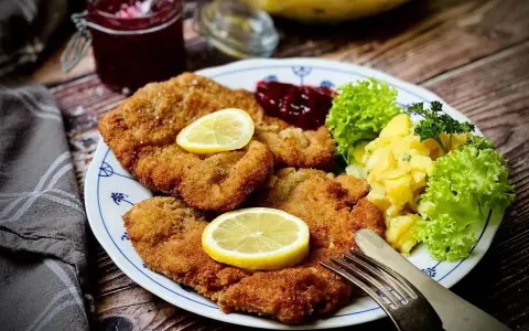 Knuspriges Wiener Schnitzel mit Zitrone, Preiselbeeren und Kartoffelsalat auf einem Teller.