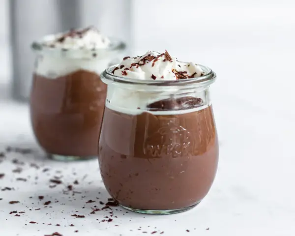 Zwei Gläser mit cremigem Schokopudding, gekrönt mit Schlagsahne und Schokostreuseln. Perfekt, um Schokopudding selber machen zu lernen!