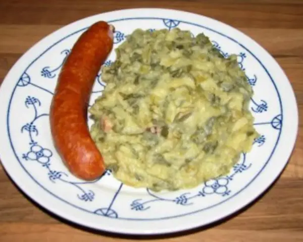Omas Stielmus Rezept: deftiger Grünkohl mit Kartoffeln und einer herzhaften Bratwurst auf einem blau-weißen Teller.