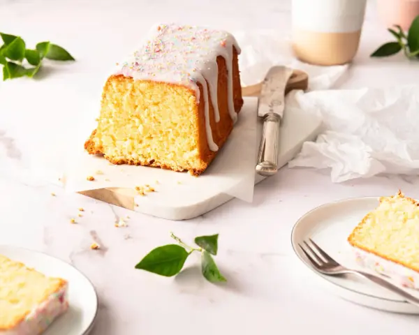 Saftiger zitronenkuchen kastenform mit Zuckerguss und bunten Streuseln, auf einem Marmorsockel, daneben ein Messer und ein Teller mit einem Stück Kuchen.