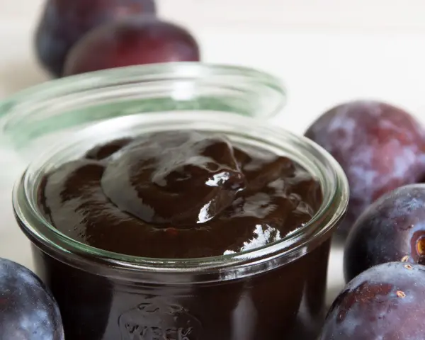 Ein Glas mit dunklem Pflaumenmus, umgeben von frischen Pflaumen. Perfekt für ein einfaches Pflaumenmus Rezept.