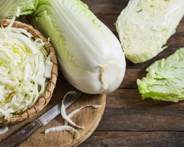 Frischer Chinakohl, bereit für leckere Chinakohl Rezepte. Ein ganzer Kohlkopf, geschnittene Blätter und fein gehackter Kohl in einem Korb liegen auf einem Holztisch.