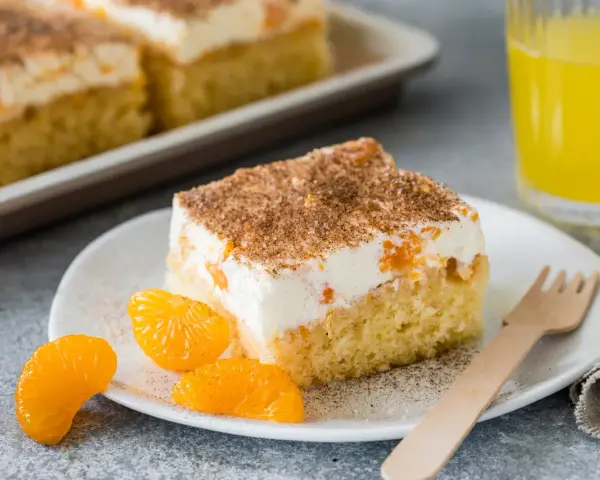 Ein saftiges Stück Fantakuchen-Rezept mit Mandarinen und Sahne, bestreut mit Zimt. Dazu ein Glas Fanta und frische Mandarinenstücke.