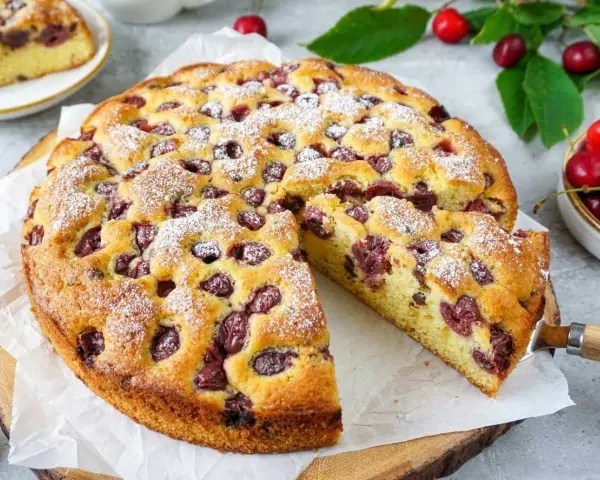 Ein saftiger Kirschkuchen, bestäubt mit Puderzucker, mit einem Stück herausgeschnitten. Frische Kirschen und Blätter liegen daneben.