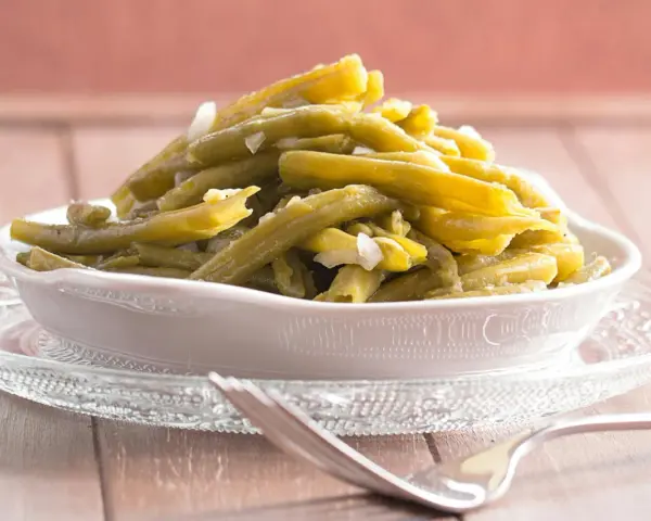 Omas Bohnensalat Rezept: Grüne Bohnen mit Zwiebeln in einer Schüssel auf einem Glasuntersetzer, daneben eine Gabel.