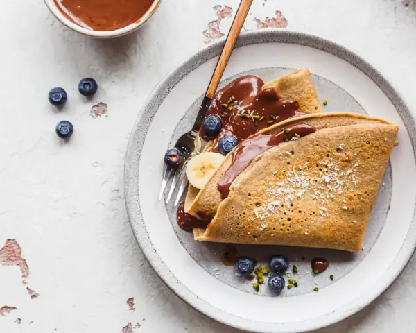 Buchweizen-Crêpes mit Schokoladensoße, Blaubeeren und Banane. Ein köstliches Beispiel für Buchweizen Rezepte, garniert mit Kokosraspeln und Pistazien.