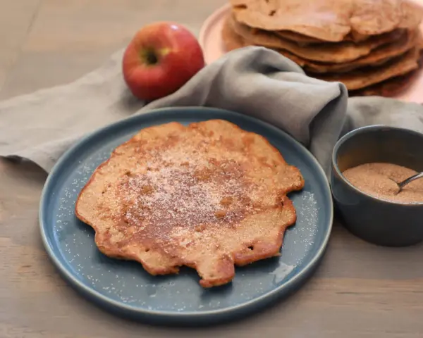 Ein Apfelpfannkuchen, bestreut mit Zucker und Zimt, liegt auf einem blauen Teller. Daneben ein roter Apfel und eine Schüssel mit Zimtzucker. Ein leckeres Apfelpfannkuchen Rezept zum Nachmachen.