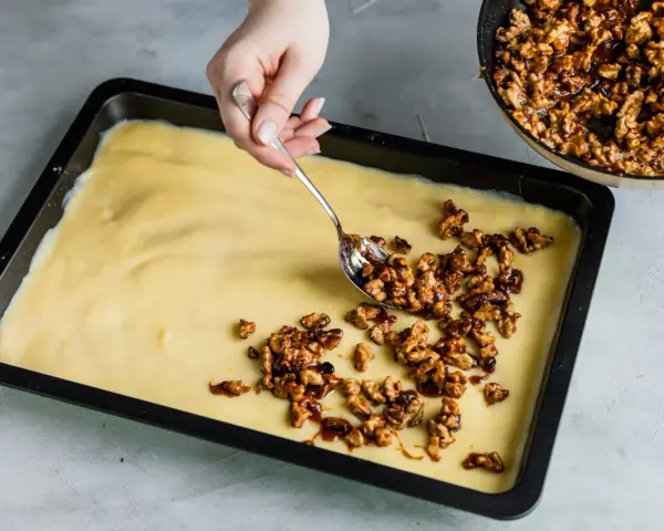 Eine Hand verteilt karamellisierte Walnüsse auf einem hellgelben Teig in einer Backform. Bald gibt es einen "friss dich dumm Kuchen"!