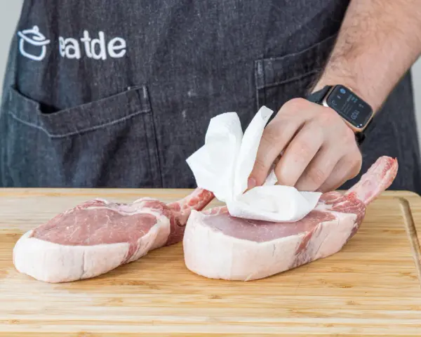Vorbereitung für ein saftiges Kotelett Rezept: Frische Schweinekoteletts werden auf einem Holzbrett mit Küchenpapier abgetupft.