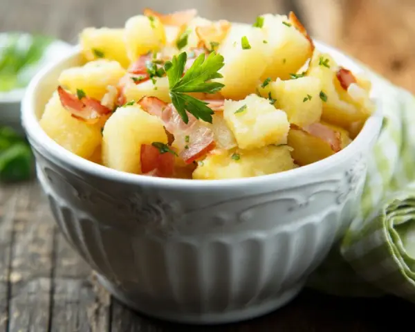 Ein einfacher Kartoffelsalat mit Speck und Petersilie in einer Schüssel, serviert auf einem Holztisch.