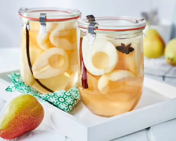 Zwei Gläser mit eingekochten Birnen, verfeinert mit Zimt und Sternanis. Ein frische Birne liegt daneben.