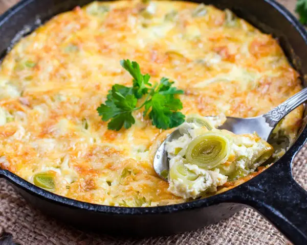 Ein goldbraunes Kartoffel-Lauch-Gratin in einer Gusseisenpfanne, mit einem Löffel herausgenommen, garniert mit Petersilie.