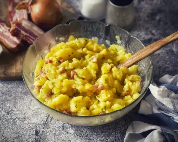Schüssel mit warmem Kartoffelsalat, garniert mit Speck und Kräutern, daneben Zwiebeln und Speck.