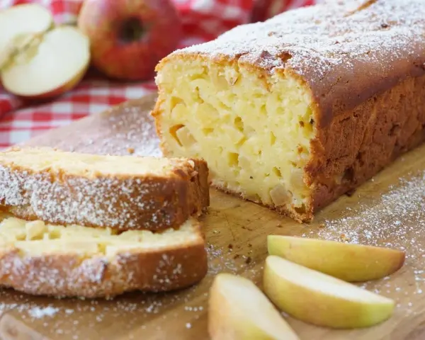 Ein saftiger, schneller Apfelkuchen mit Puderzucker bestäubt, daneben Apfelspalten und ganze Äpfel auf kariertem Tuch.