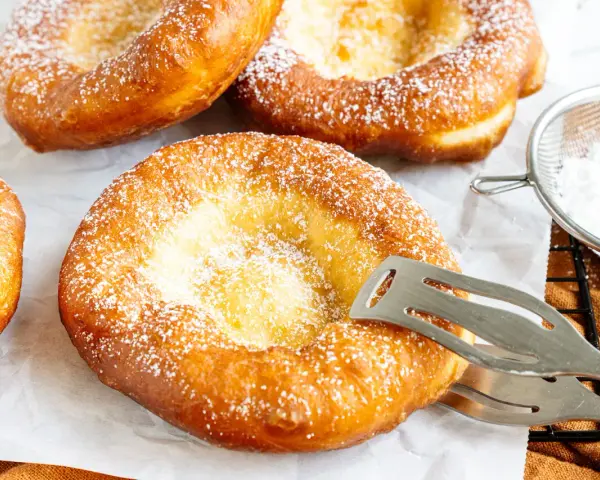 Frisch gebackene, ausgezogene Krapfen, bestäubt mit Puderzucker. Ein Kochlöffel hält einen der goldbraunen Krapfen.
